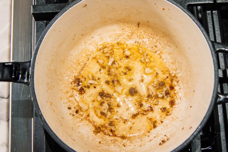 Chopped garlic and onions are sautéing in oil at the bottom of a white pot on a stovetop, with the mixture starting to brown and become fragrant.
