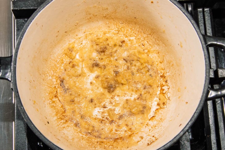 A close-up of a pot on a stovetop shows browned flour and fat cooking together, forming a roux with a light brown color and bubbly texture.