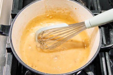 A whisk stirs a creamy, light brown sauce with visible bits in a white pot on a stovetop.