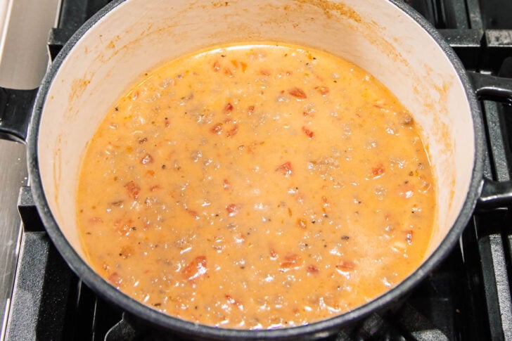 A large white pot on a stovetop contains a creamy tomato sauce with visible chunks of tomatoes and herbs, simmering and mixed well.