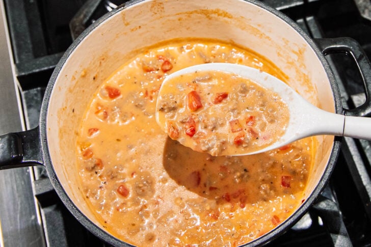 A pot of creamy, orange-colored soup with chunks of ground meat and diced tomatoes, being stirred and scooped with a white ladle on a stovetop.
