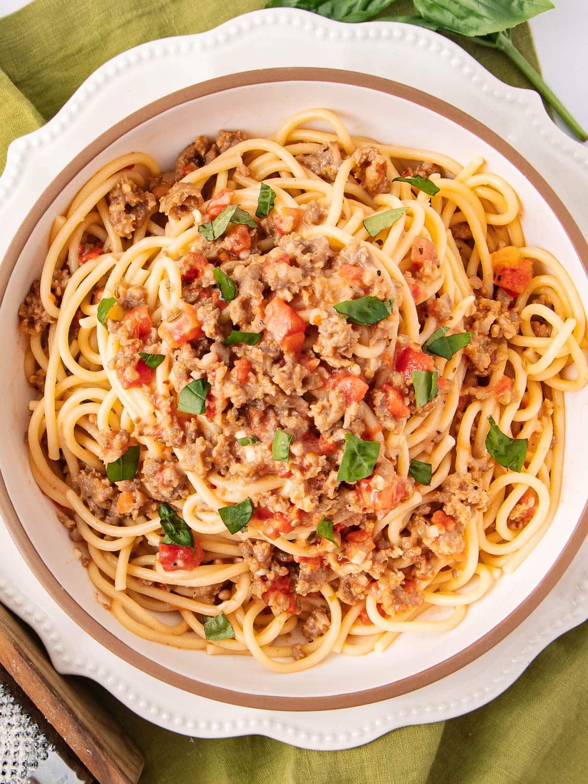 A white bowl filled with spaghetti topped with a chunky meat and tomato sauce, garnished with chopped fresh basil, sits on a green cloth.