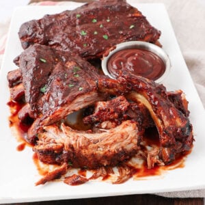 A plate of saucy, cooked pork ribs garnished with herbs, served with a small metal cup of barbecue sauce on the side. The ribs are tender, with some pieces pulled apart to show the juicy meat inside.