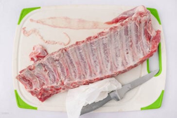 A slab of raw pork ribs on a white cutting board with green edges, alongside a knife partially wrapped in a paper towel and some loose meat and membrane scraps.