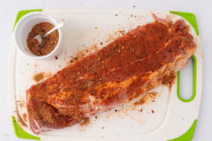 A large raw pork tenderloin coated with spices sits on a white cutting board with green edges. A small white bowl filled with spice mix and a spoon is beside the meat.