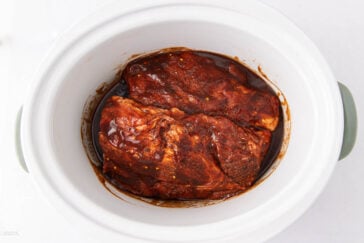 Raw, marinated pork coated in barbecue sauce sits in a white slow cooker, ready to be cooked.