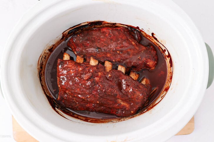 Slow-cooked pork ribs coated in barbecue sauce inside a white crockpot, with sauce pooling at the bottom. The ribs are tender and browned, partially exposing the rib bones.