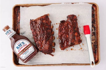 A baking sheet with parchment paper holds two sauced racks of ribs, a bottle of Ray’s Sweet & Smoky Barbecue Sauce, and a red-tipped basting brush.