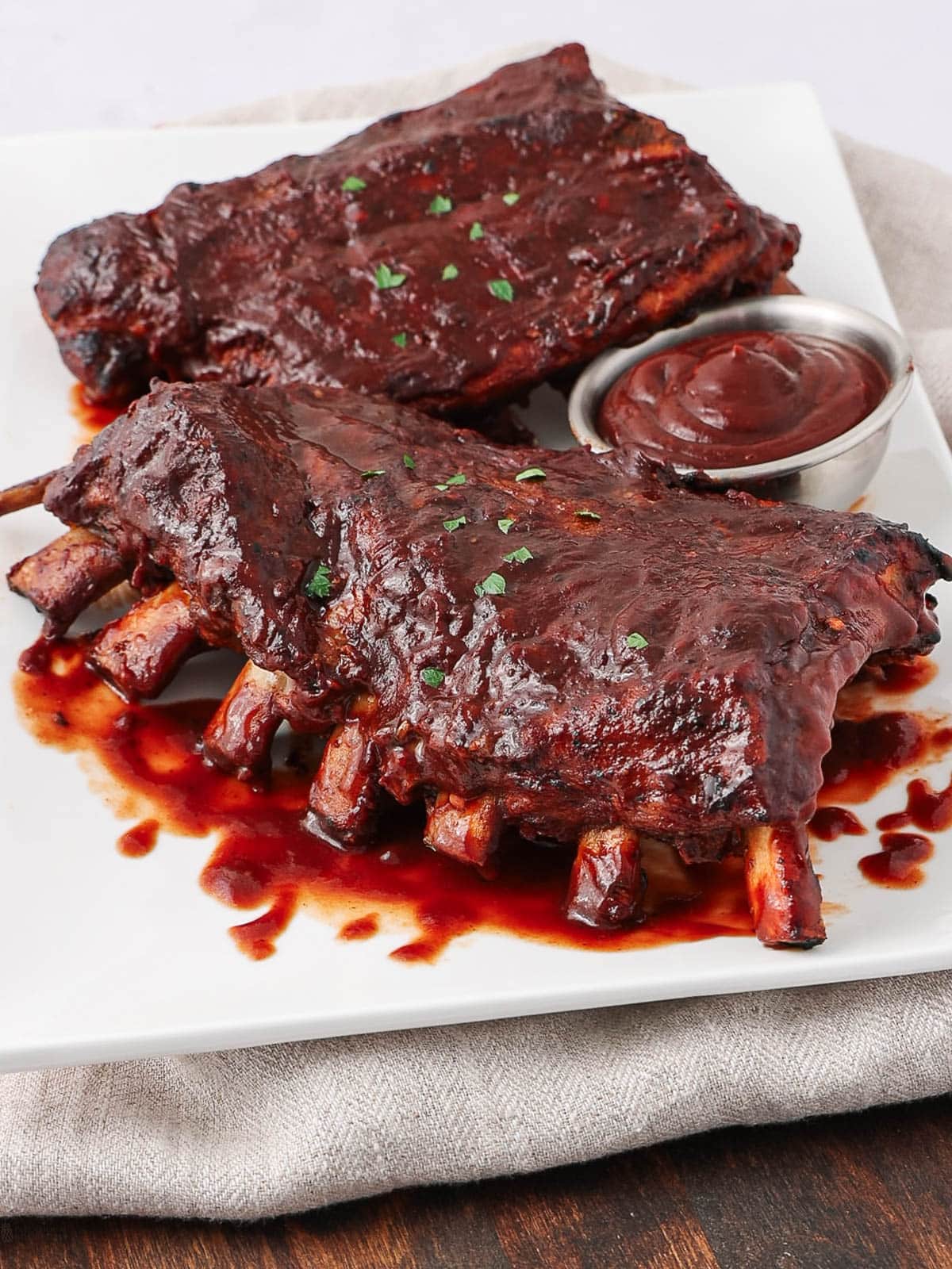 Two racks of barbecued ribs with a glossy sauce sit on a white plate, garnished with chopped herbs, next to a small dish of barbecue sauce. The plate rests on a light-colored napkin.