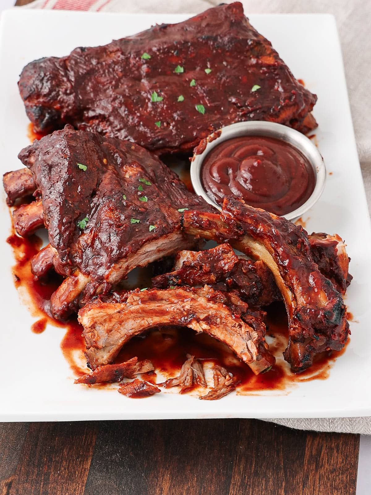 A white plate with barbecued pork ribs covered in sauce, some ribs cut and arranged around a small metal cup filled with barbecue sauce. The ribs are garnished with a sprinkle of chopped herbs.