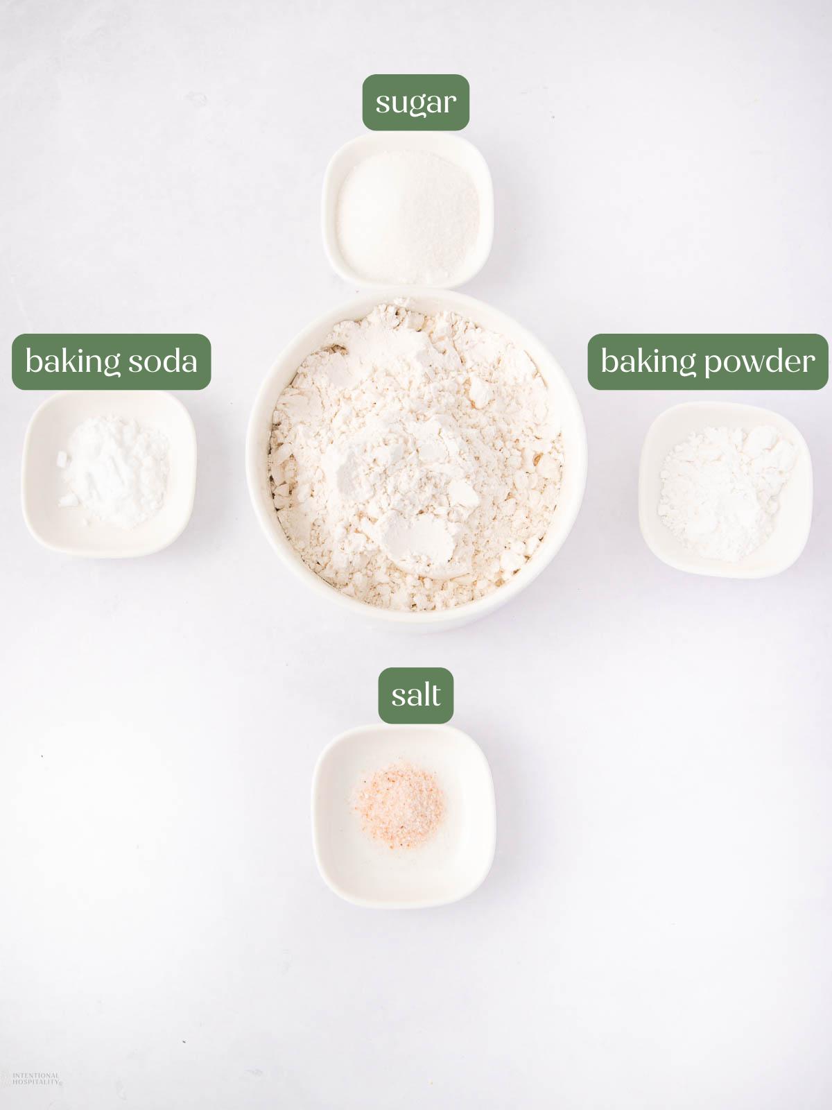 Five small white bowls on a white surface hold flour, sugar, baking soda, baking powder, and salt. Each bowl is labeled with green text above it indicating its contents.
