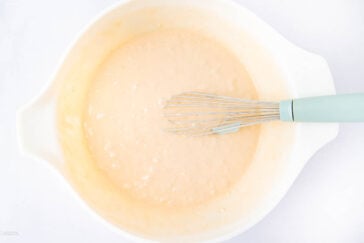 A light blue-handled whisk rests in a white mixing bowl filled with pale, slightly lumpy batter on a white surface.