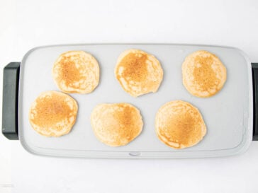 Six golden brown pancakes cooking on a rectangular electric griddle with a white background.