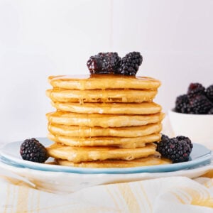A stack of pancakes topped with blackberries and drizzled with syrup sits on a white plate, with additional blackberries in a small bowl in the background.
