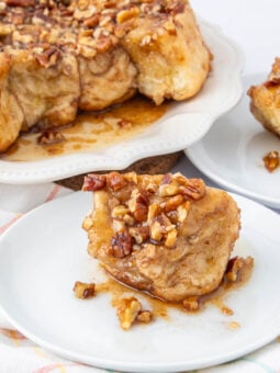 A piece of sticky pecan cinnamon monkey bread sits on a white plate, topped with chopped pecans and caramel glaze. In the background, more monkey bread is on a larger serving plate.