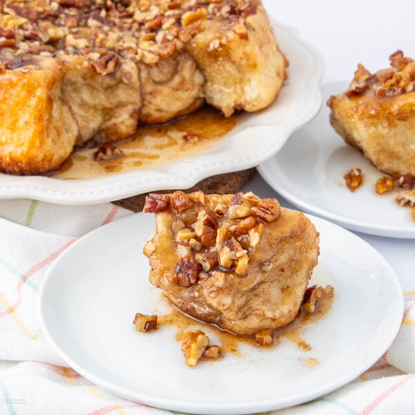 A piece of sticky pecan cinnamon monkey bread sits on a white plate, topped with chopped pecans and caramel glaze. In the background, more monkey bread is on a larger serving plate.