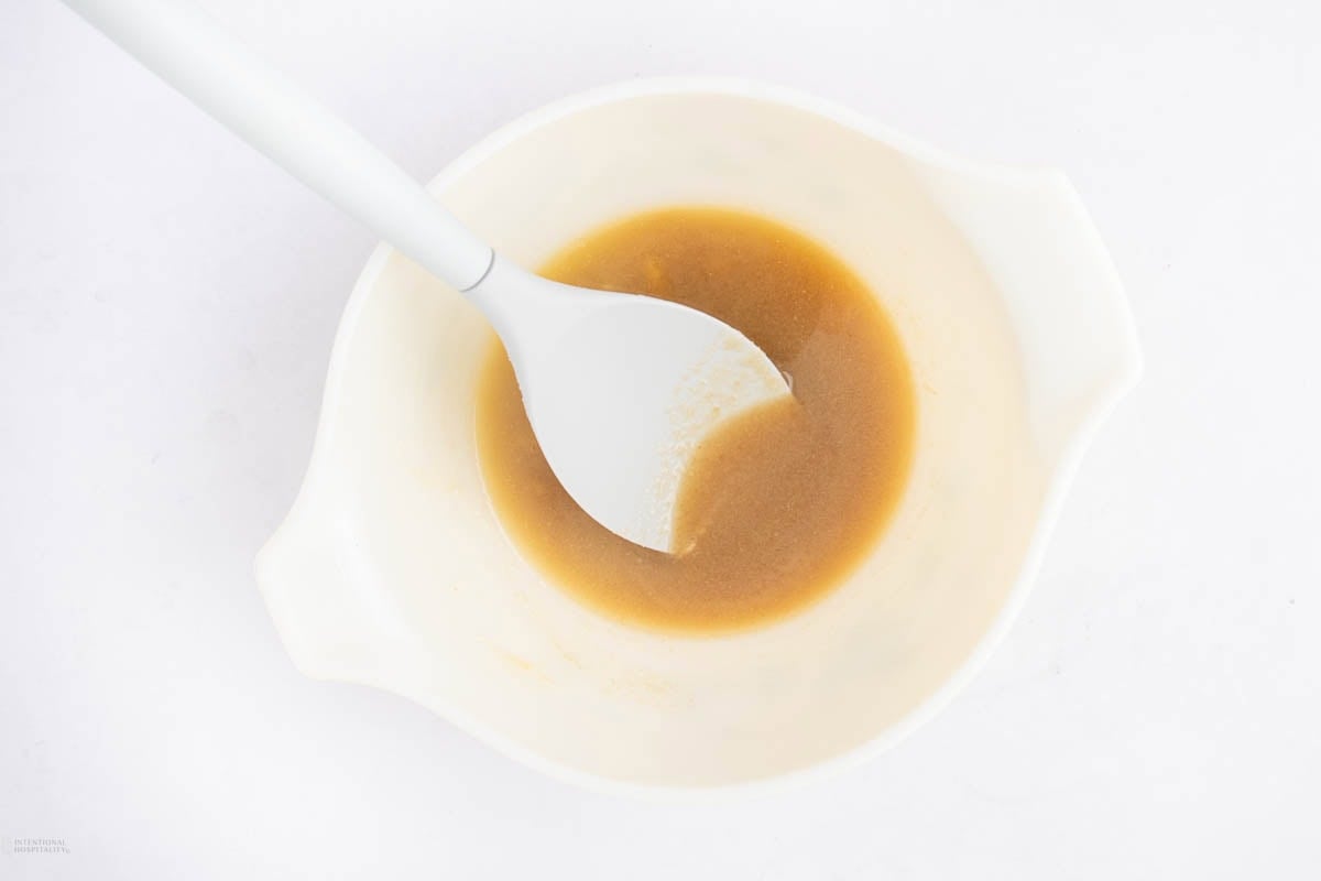 A white mixing bowl containing light brown liquid and a white spatula, viewed from above on a white surface.