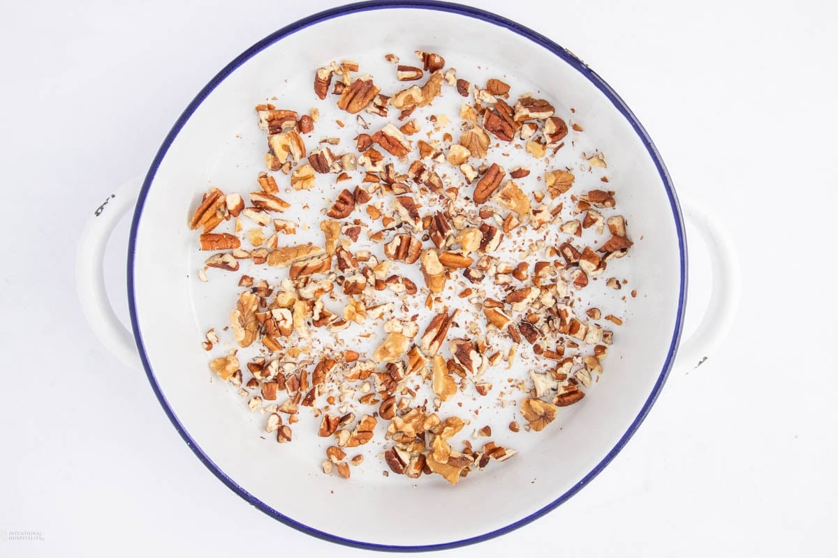 A white dish with a blue rim contains a scattered layer of chopped pecans on a white background.
