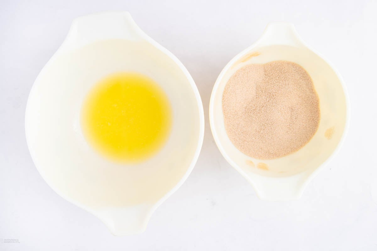 Two white mixing bowls on a white surface; the left bowl contains melted butter, and the right bowl contains granulated brown sugar.
