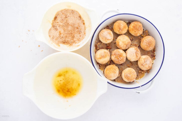Overhead view of dough balls coated in cinnamon sugar in a round baking dish, surrounded by two white bowls—one with melted butter and the other with cinnamon sugar.