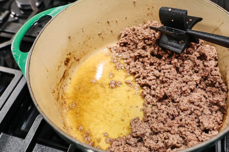 Ground beef is being browned in a large, green Dutch oven on a stovetop. A black spatula rests in the pot, separating cooked beef from rendered fat and juices pooling on one side.