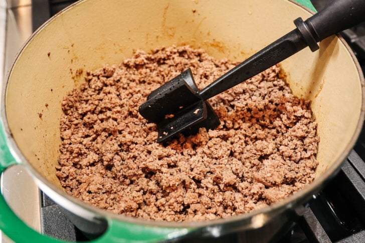 A pot filled with browned ground beef on a stovetop, being stirred with a black spatula.