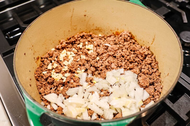 Ground beef cooking in a large pot on a stovetop, with chopped onions and minced garlic added but not yet mixed in.