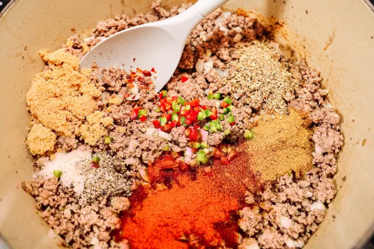Ground meat in a pot with diced green and red peppers, chopped onions, and various spices being mixed with a white spoon, ready to be cooked.