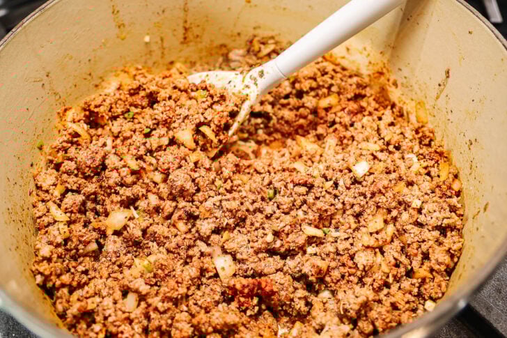 Ground beef cooked with chopped onions and spices in a large pot, being stirred with a white-handled spoon. The mixture appears well-seasoned and crumbled, ready for use in a recipe.