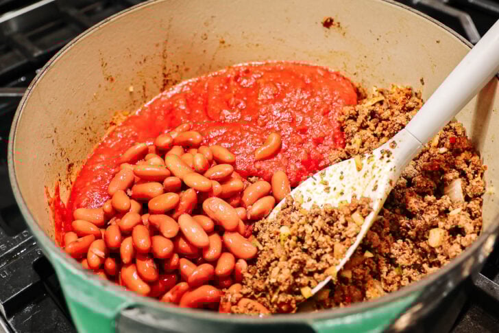A pot on a stovetop filled with ground beef, red kidney beans, and tomato sauce being stirred together with a white spatula, preparing a chili dish.