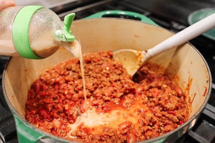 A hand pours a creamy liquid from a green-lidded bottle into a pot of chili with ground meat, beans, and tomato sauce, while a white spoon rests in the pot.