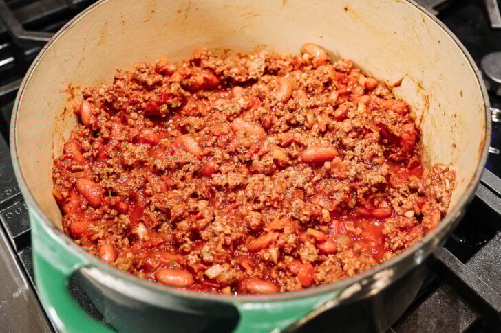 A large pot filled with homemade chili made of ground beef, kidney beans, and tomato sauce cooks on a stovetop.