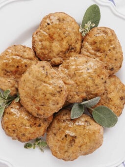 A white plate with eight golden-brown sausage patties, garnished with fresh green herbs and small white flowers, on a white background.