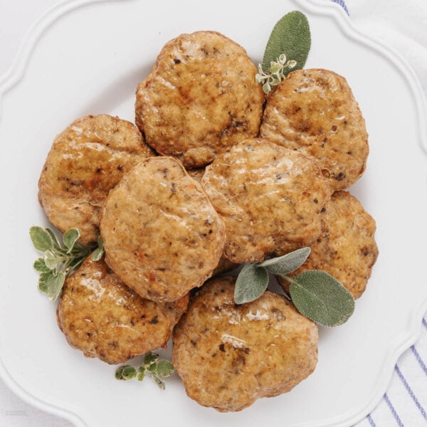 A white plate with eight golden-brown sausage patties, garnished with fresh green herbs and small white flowers, on a white background.