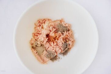 Raw ground meat with various spices, including black pepper, paprika, and possibly garlic or onion powder, placed on top in a white mixing bowl on a plain white background.
