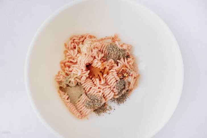 Raw ground meat with various spices, including black pepper, paprika, and possibly garlic or onion powder, placed on top in a white mixing bowl on a plain white background.