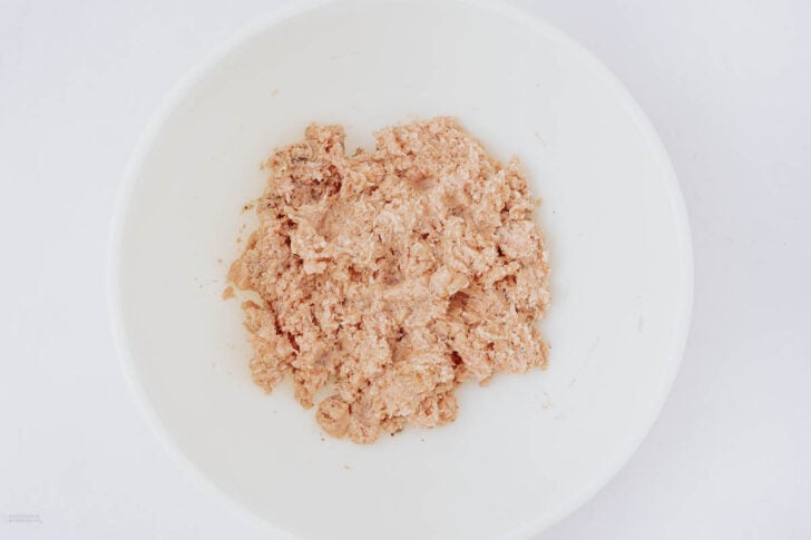 A white bowl containing a mixture of light brown, finely mashed or blended food, possibly tuna or another type of meat spread, set against a plain white background.