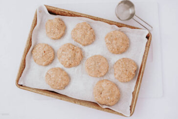 A baking tray lined with parchment paper holds nine unbaked dough balls, spaced apart. A metal measuring cup rests nearby on a white surface.