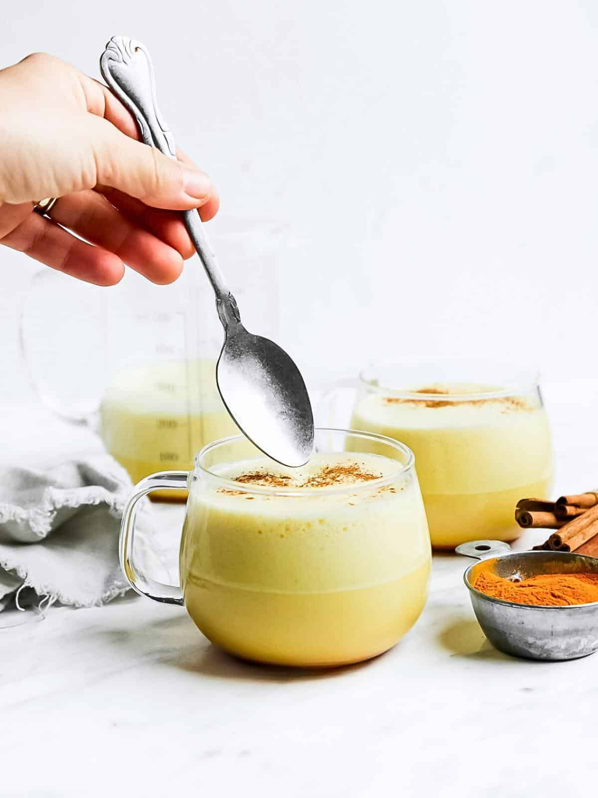 A hand holds a spoon above a glass mug filled with a frothy yellow drink, likely golden milk, with two similar mugs, cinnamon sticks, and a bowl of turmeric powder nearby on a white surface.