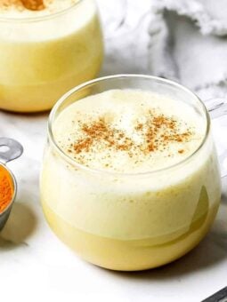 A clear glass mug filled with golden turmeric latte topped with frothy foam and sprinkled with cinnamon, next to a small metal cup of turmeric powder and cinnamon sticks on a white surface.