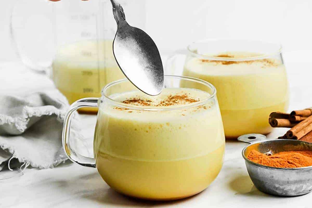A hand holds a spoon above a clear glass mug filled with a frothy golden drink, topped with a sprinkle of cinnamon. Two more mugs, whole cinnamon sticks, and a small bowl of ground turmeric are in the background.