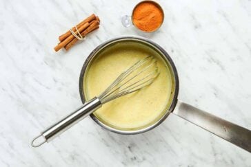 A saucepan filled with a creamy yellow mixture being whisked, next to a small bundle of cinnamon sticks and a metal cup of ground cinnamon, all on a white marble surface.