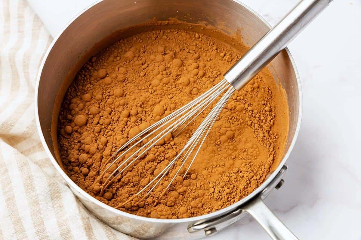 A metal saucepan filled with a layer of cocoa powder being whisked with a metal whisk, sitting on a white surface next to a striped cloth.