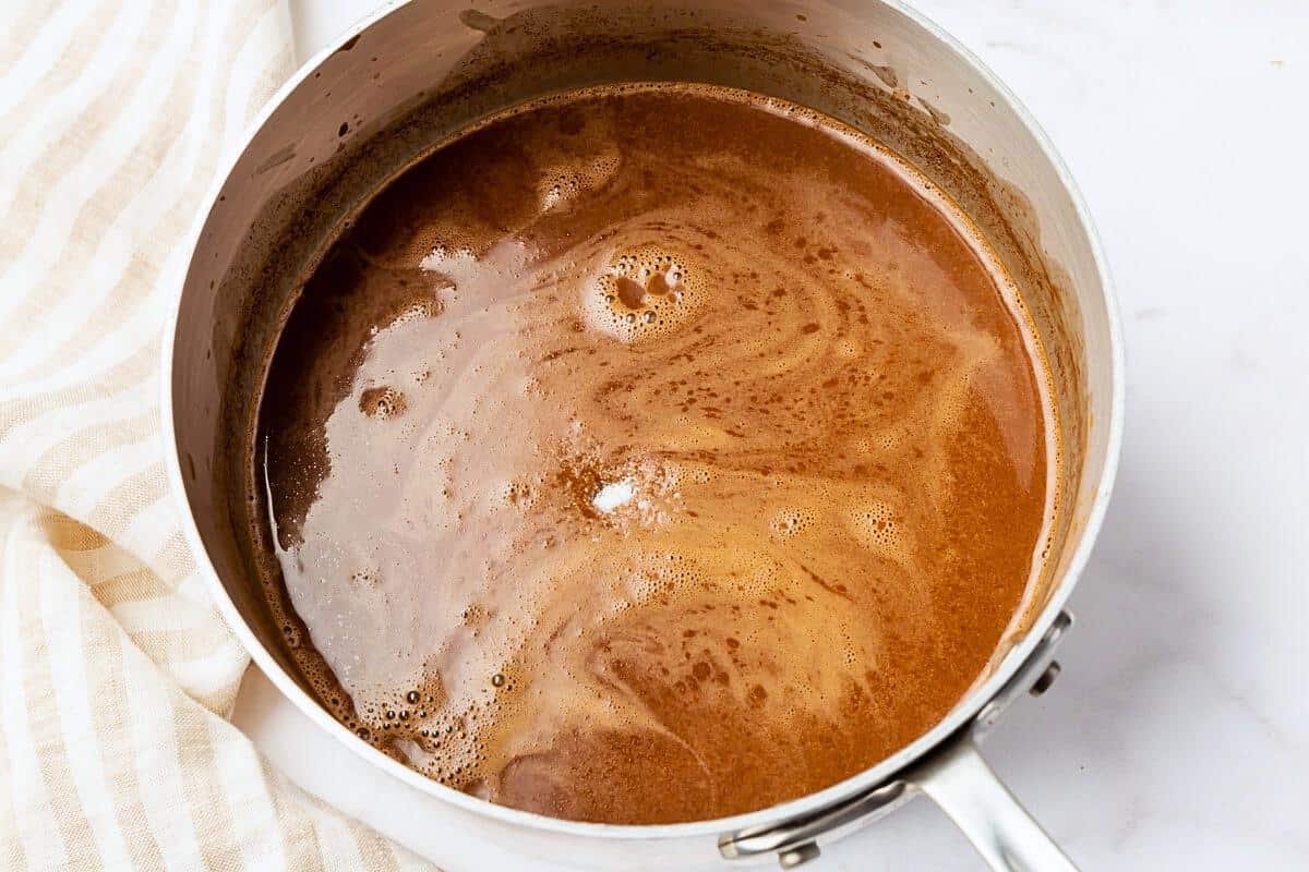 A silver saucepan filled with brown chicken broth sits on a white surface next to a beige striped cloth.