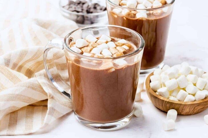 Two glass mugs of hot chocolate topped with mini marshmallows sit on a light surface next to a wooden bowl of more marshmallows, a small bowl of chocolate chips, and a beige striped cloth.