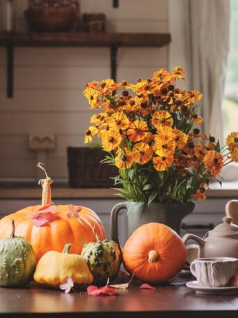 A vase of orange flowers, pumpkins, and gourds are arranged on a kitchen table with a teapot, cup, and autumn leaves. The background shows shelves and a window with sunlight streaming in.