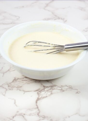 A metal whisk rests in a white bowl filled with smooth, creamy batter on a white marble countertop.