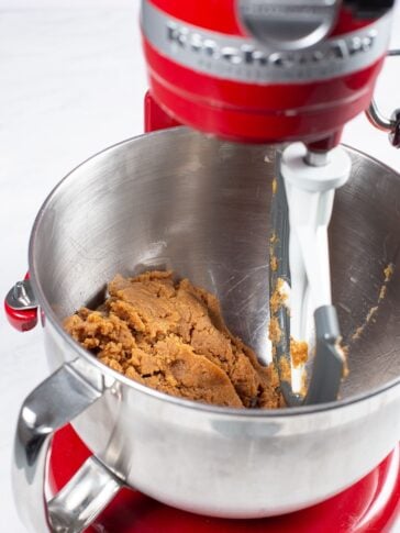 A red stand mixer with a paddle attachment is mixing brown cookie dough in a stainless steel bowl. The dough is partially combined and sticks to the sides of the bowl.