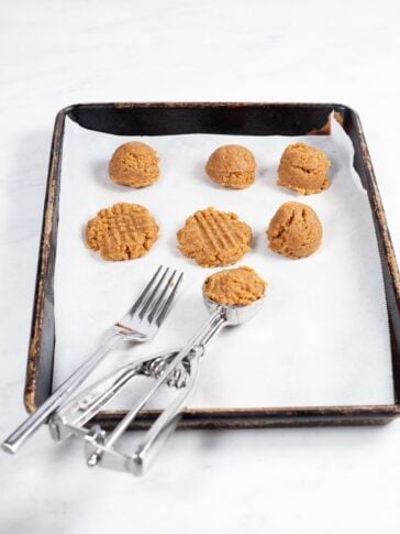 A baking tray lined with parchment paper holds scoops of peanut butter cookie dough, some with fork marks. A metal cookie scoop and two forks rest on the tray's edge.