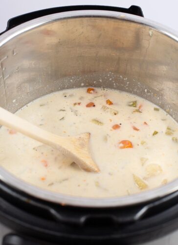 A creamy soup with chopped vegetables is being stirred with a wooden spatula inside a stainless steel Instant Pot.